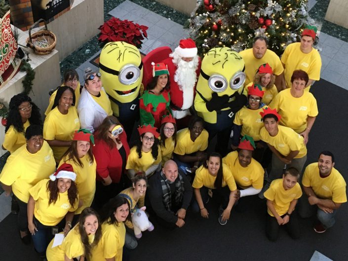 velká skupina zaměstnanců ADP ve žlutých tričkách stojící kolem Santa Clause, elfa a dvou postaviček Mimoňů z filmu Já, padouch.